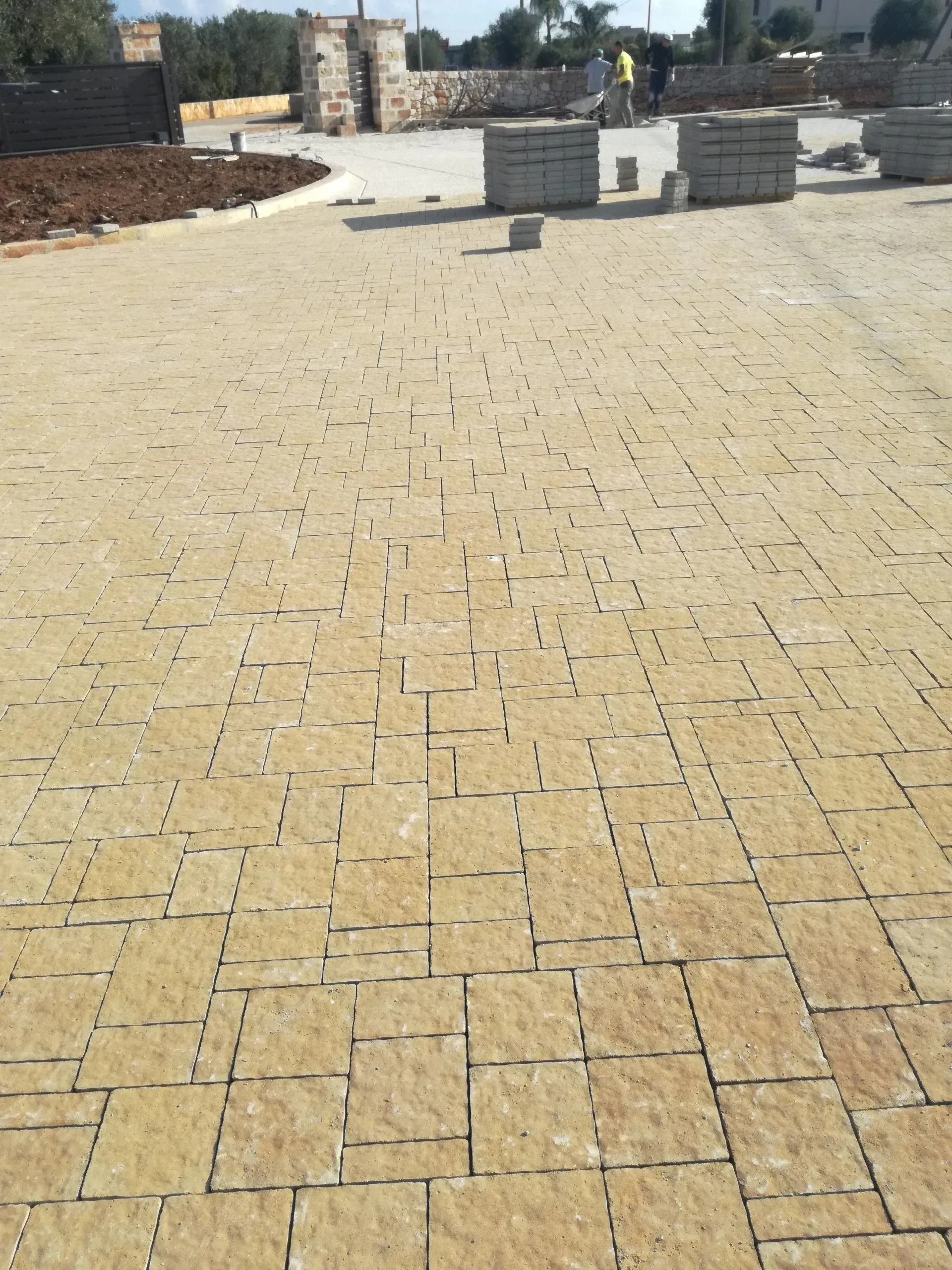 Pavimentazioni in pietra gialla in fase di posa per una piazza o un sentiero; sullo sfondo pile di pavimentatori e operai.