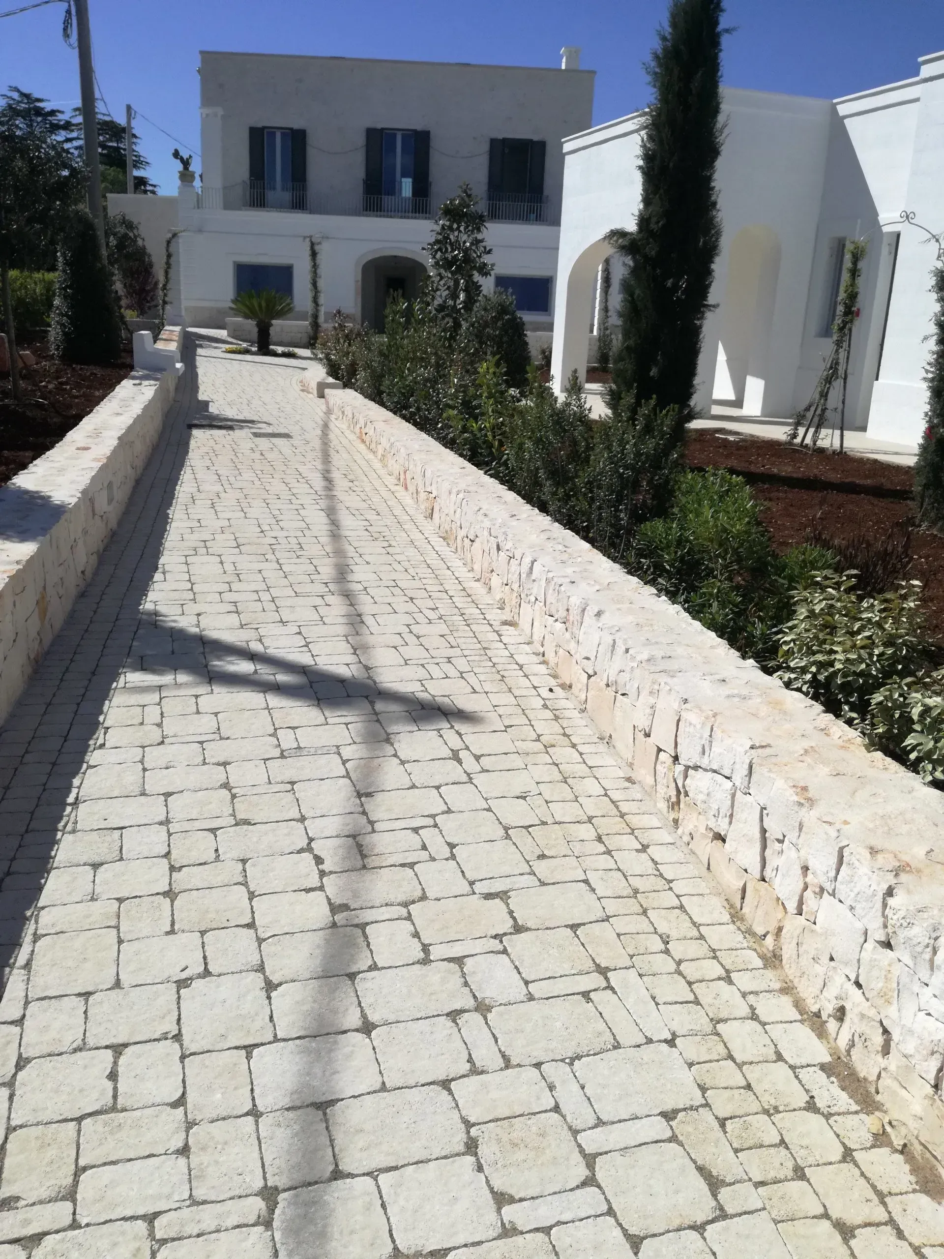 Sentiero acciottolato che conduce a un edificio bianco con finestre, delimitato da muri in pietra e vegetazione.