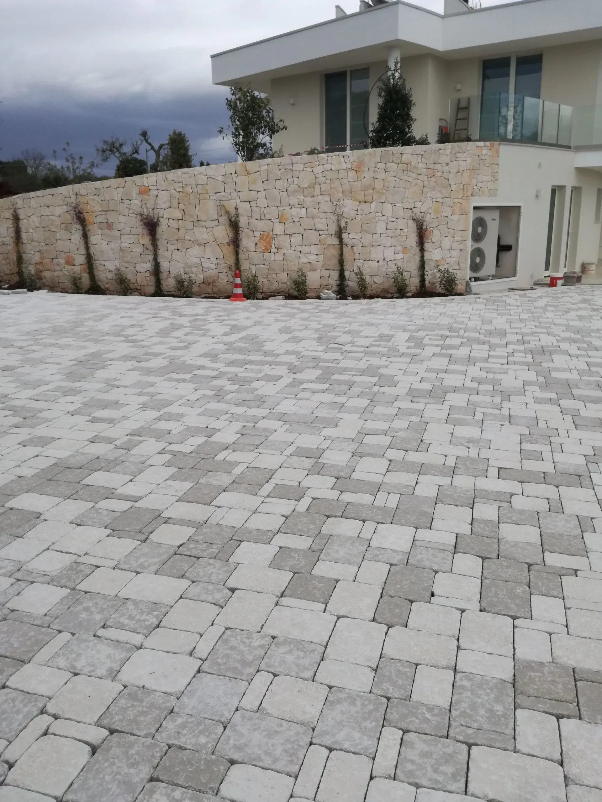 Vialetto lastricato in pietra di fronte a una casa moderna con un muro in pietra e alcune piante. Cielo nuvoloso.
