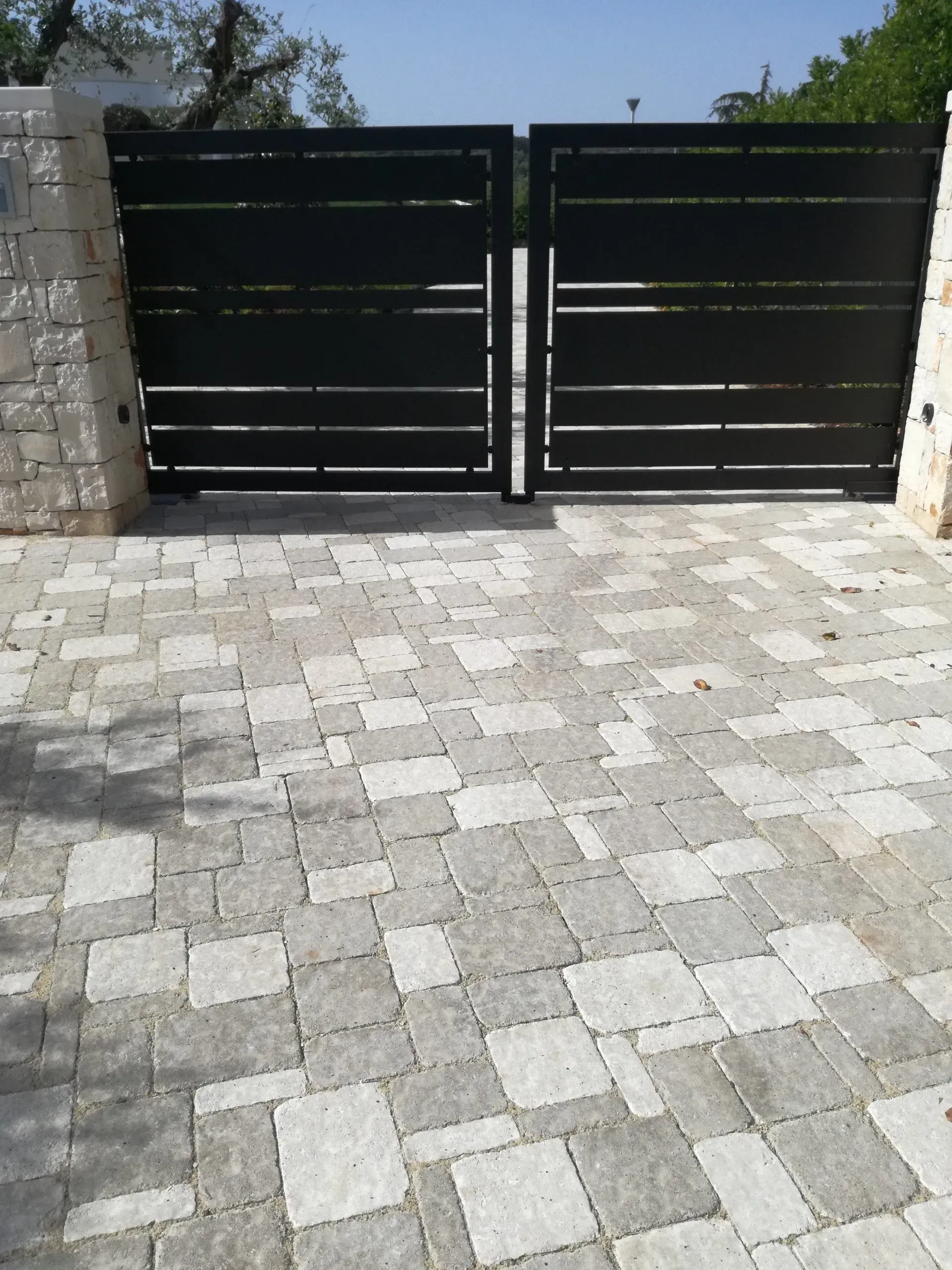 Cancello in metallo nero con sbarre orizzontali, incastonato tra colonne in pietra, su un vialetto in pietra grigia.