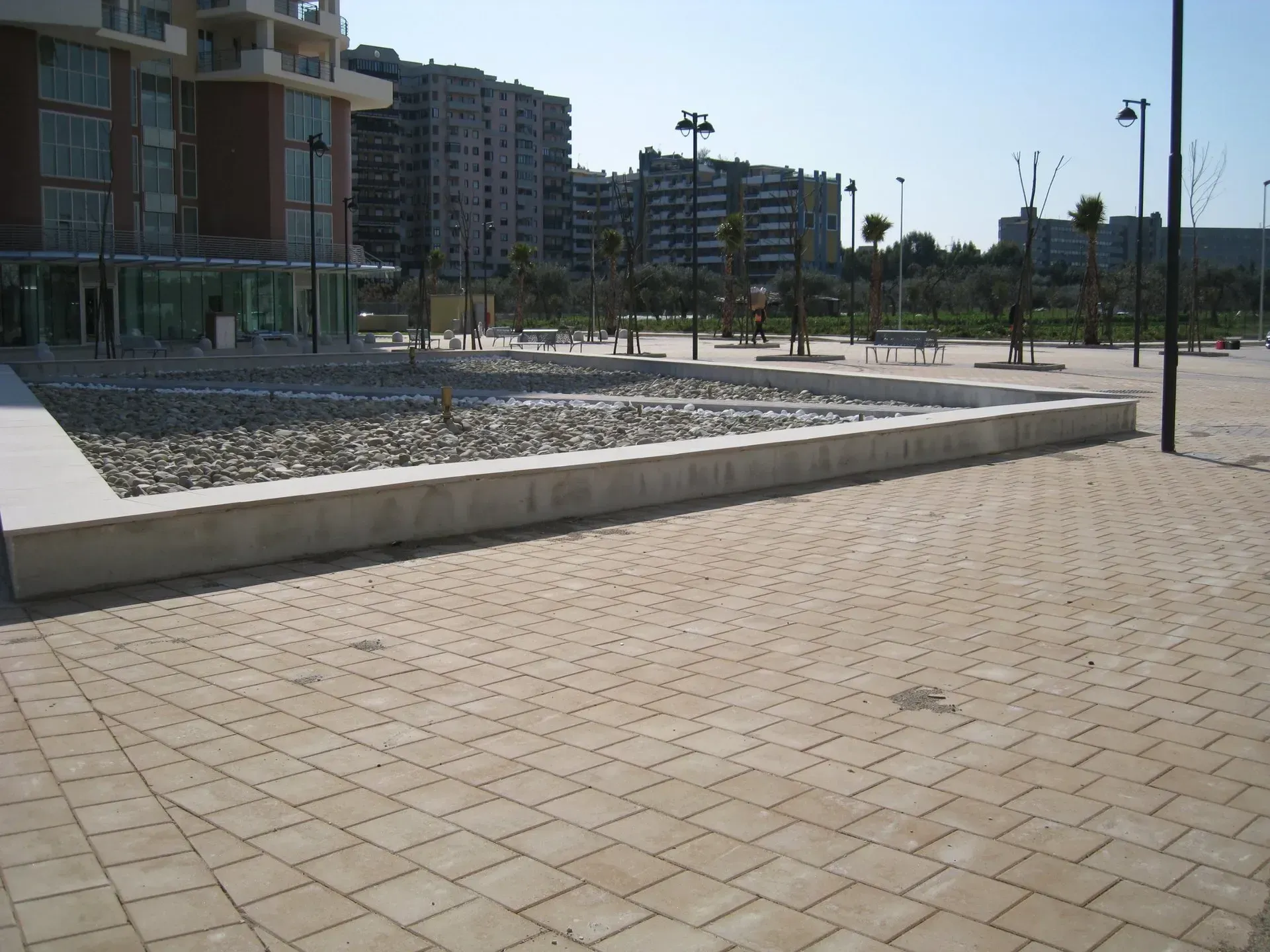 Piazza di cemento con un lungo bordo in pietra riempito di ciottoli, di fronte a un edificio moderno.