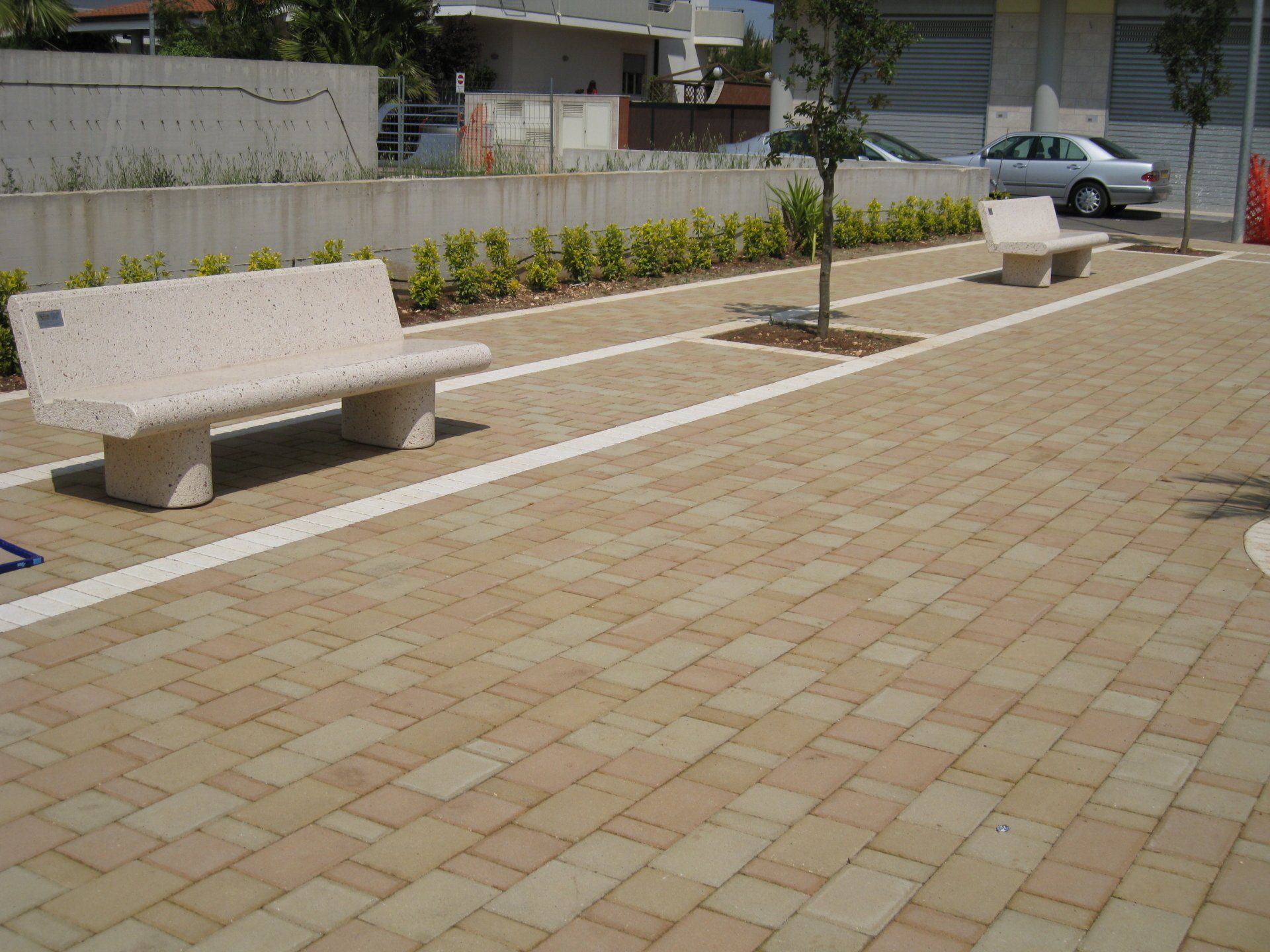 Panchine in pietra su un sentiero lastricato, fiancheggiato da un muretto con cespugli e piccoli alberi.