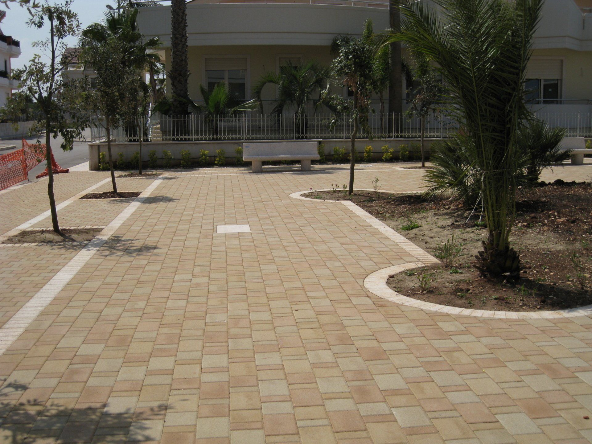 Spazio pubblico pavimentato in mattoni con alberi, una panchina e un edificio sullo sfondo in una giornata di sole.
