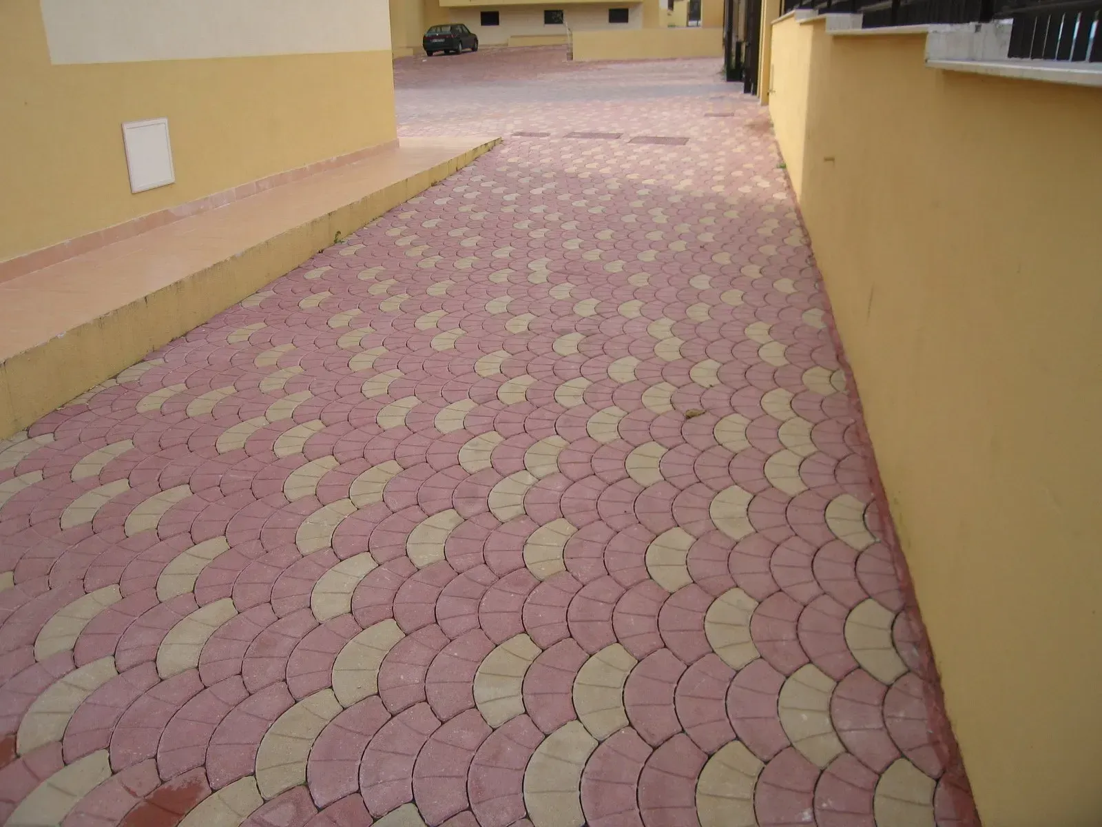 Passerella pavimentata con mattoni rossi e beige a forma di ventaglio. Muri gialli delimitano la passerella; un'auto è visibile in lontananza.