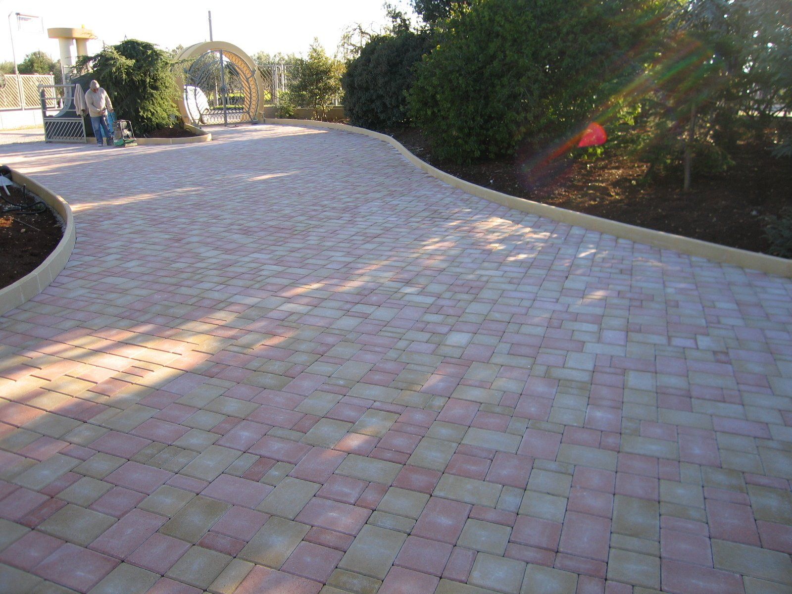 Vialetto in mattoni che curva verso un giardino alberato, con superficie chiara nelle tonalità del rosa e del beige.