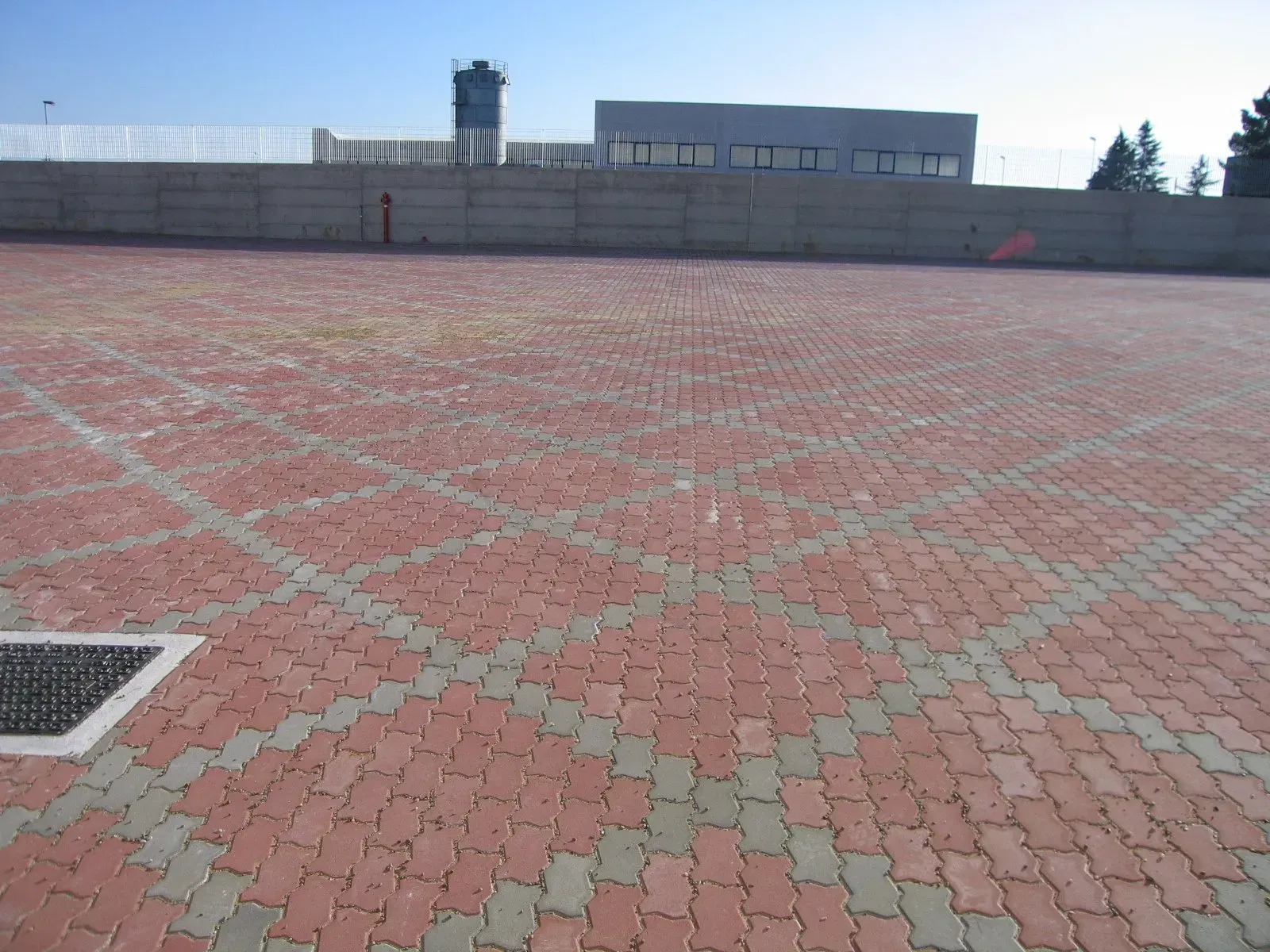 Parcheggio pavimentato in mattoni rossi e grigi con un basso muro di cemento e un edificio sullo sfondo.