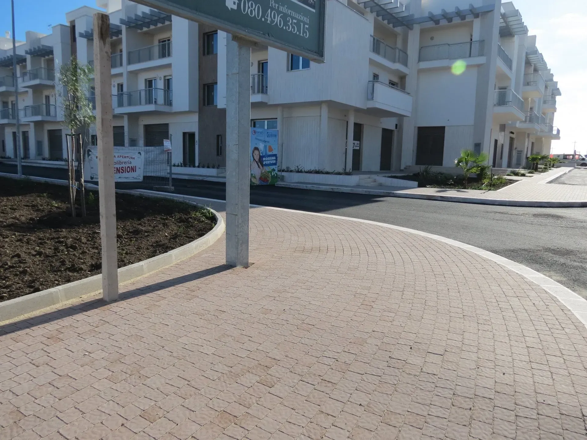 Marciapiede curvo in mattoni che conduce a un edificio con balconi. Cartello e marciapiede sulla sinistra.