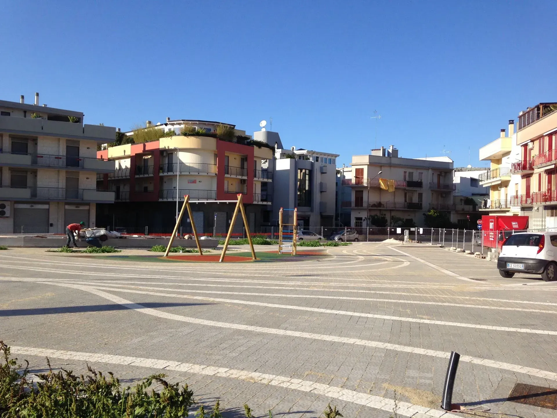 Una piazza aperta e pavimentata con un'altalena, circondata da condomini sotto un cielo azzurro.