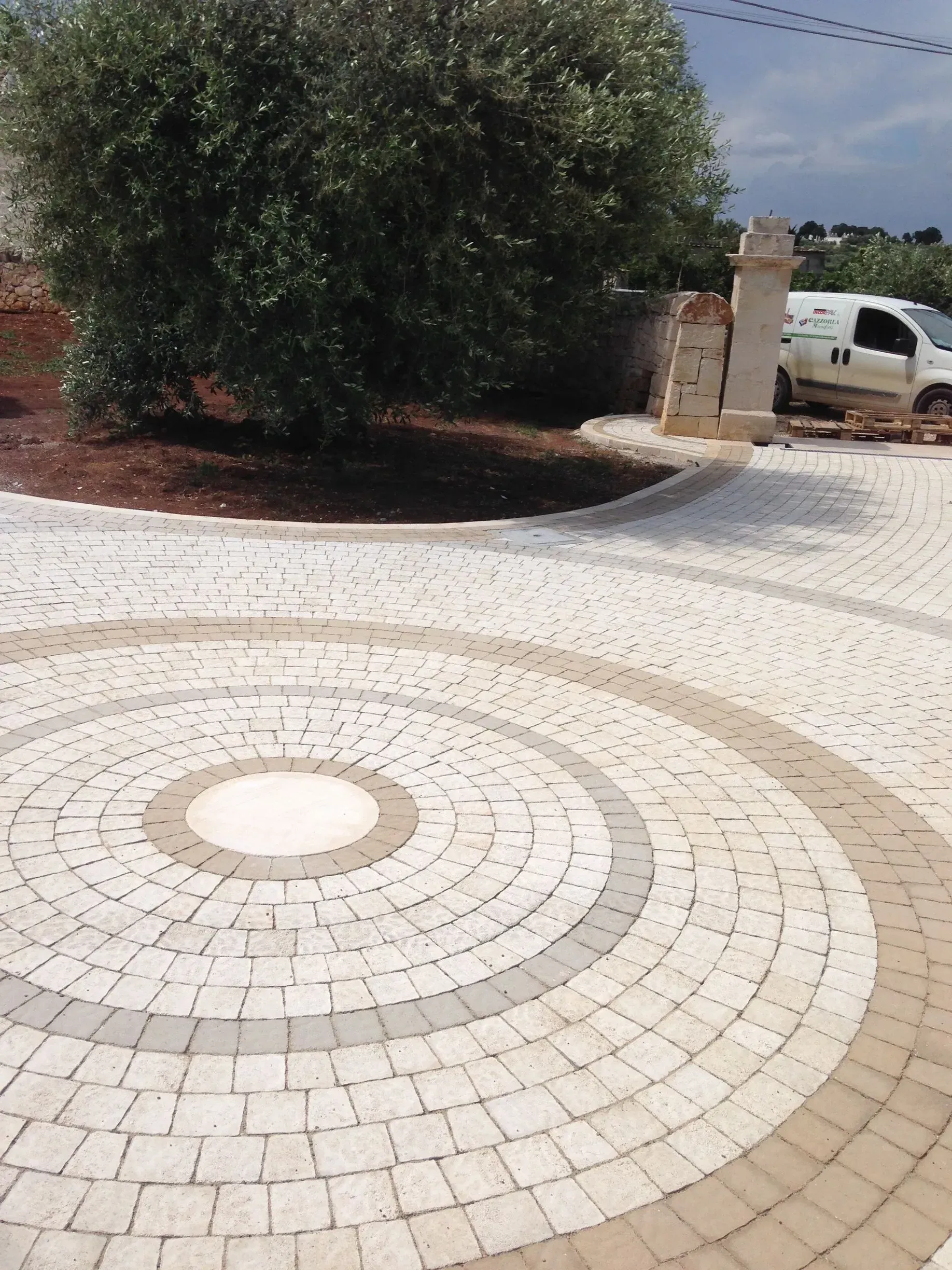 Vialetto circolare pavimentato in mattoni con ulivi, tonalità beige e grigio. Veicolo bianco visibile.