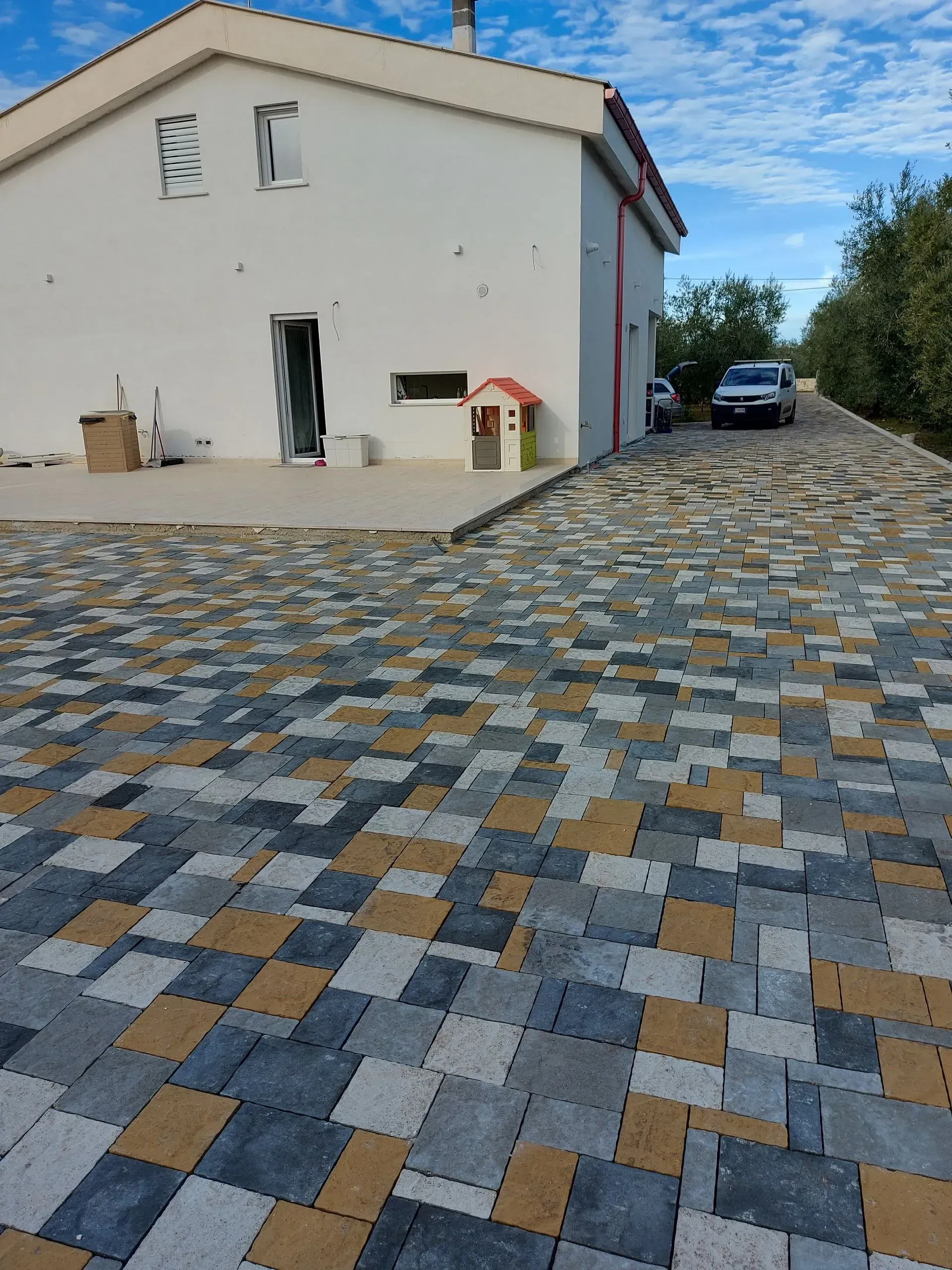 Casa bianca con vialetto pavimentato, un'auto parcheggiata. Pavimentazioni quadrate multicolori. Cielo azzurro.