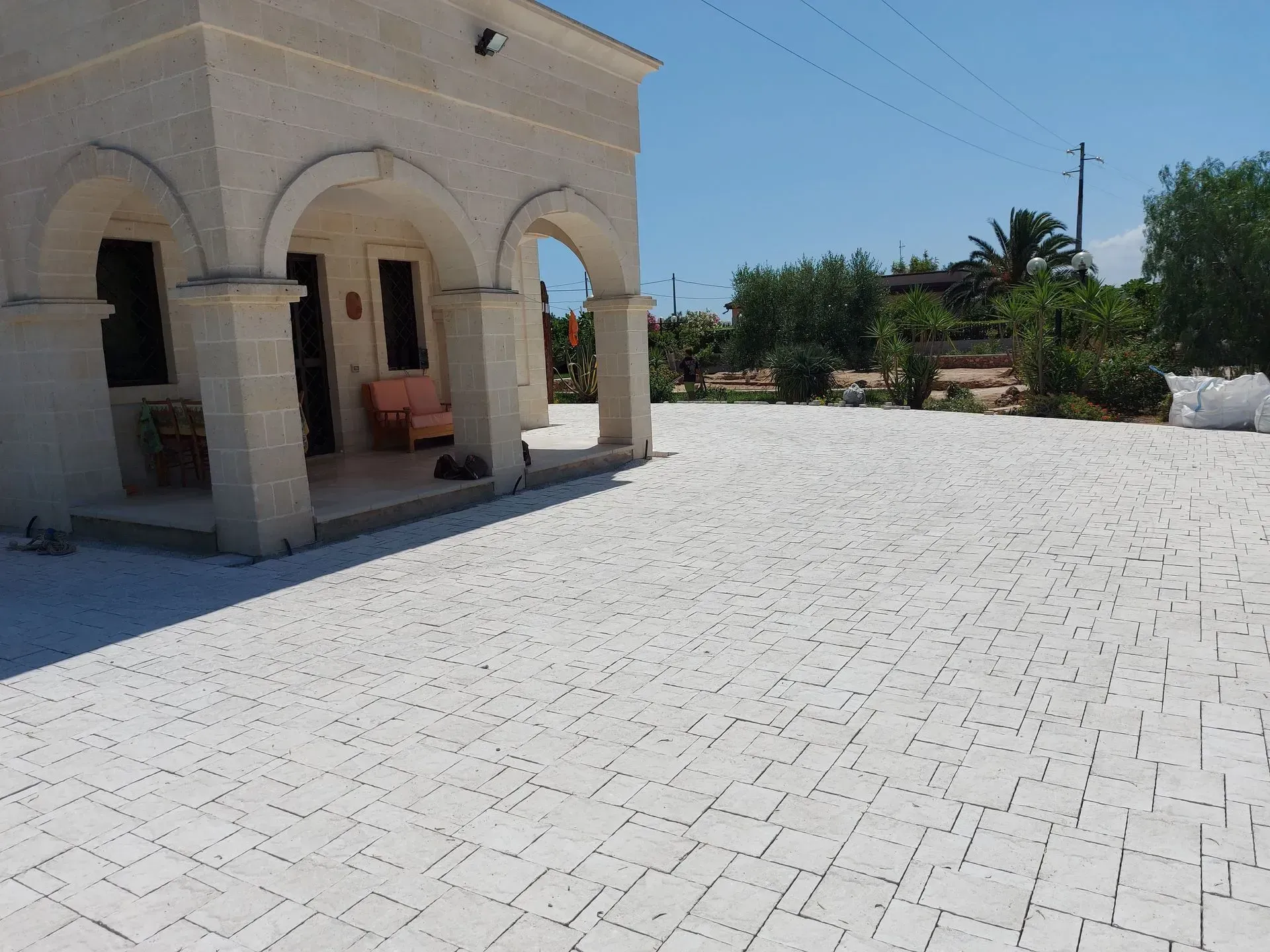 Cortile pavimentato in pietra bianca con ingresso ad arco che conduce a un edificio di colore chiaro, sotto un cielo azzurro brillante.