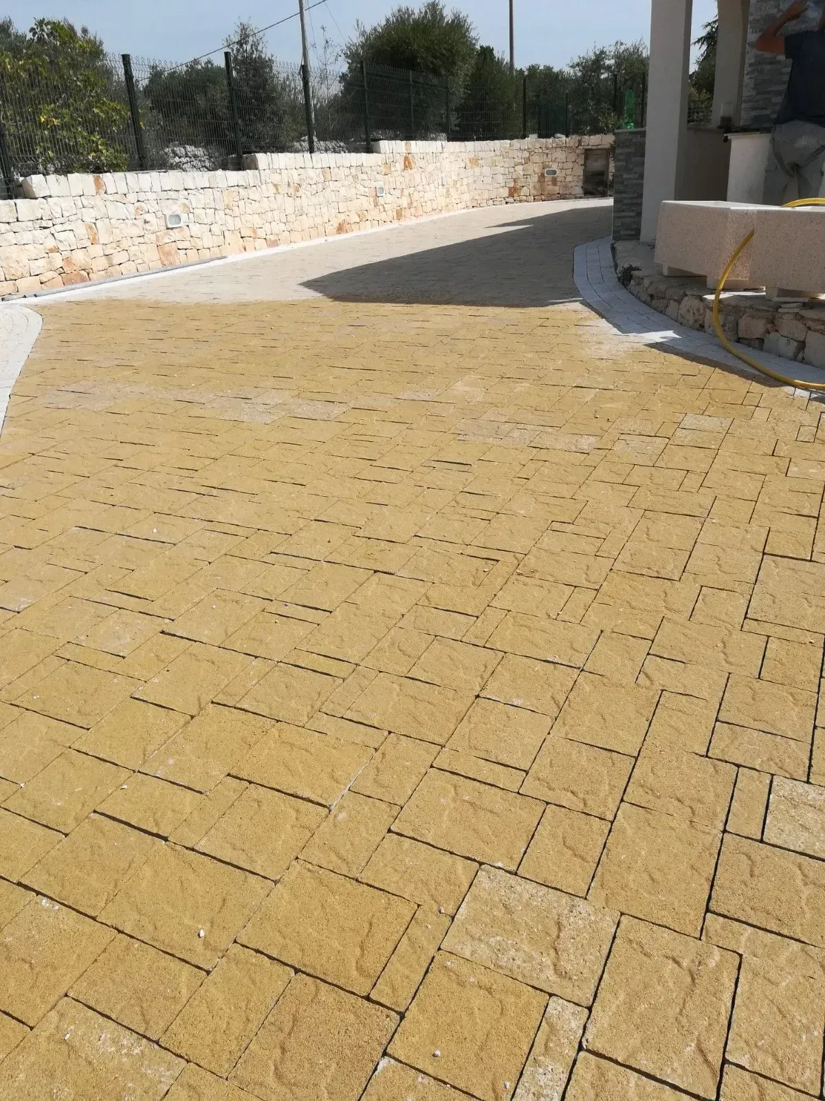 Vialetto di mattoni gialli che conduce a un edificio, delimitato da una striscia di cemento bianco e da un basso muro di pietra.