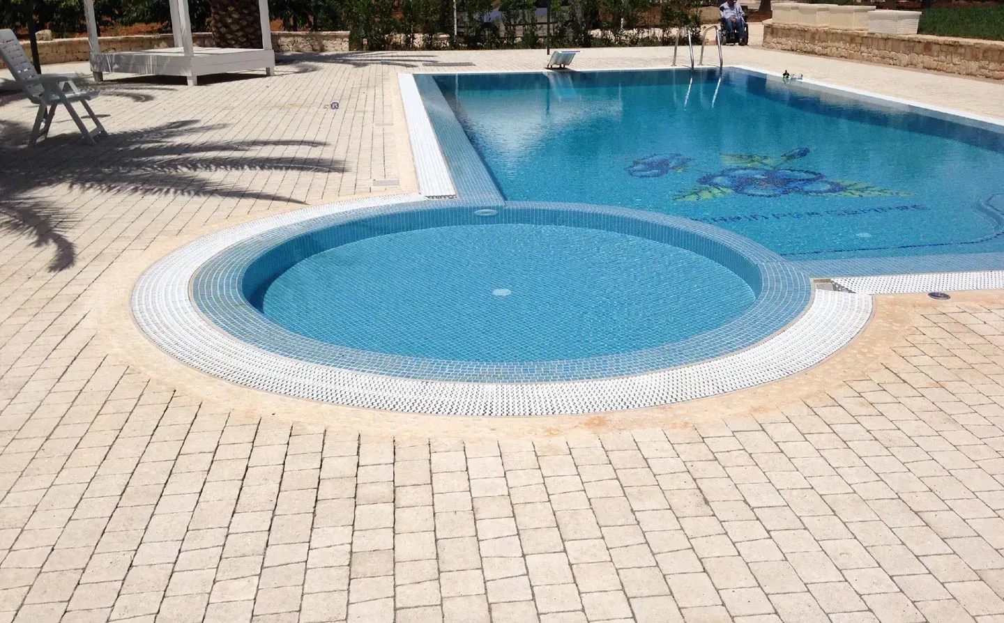 Piscina con jacuzzi circolare, circondata da pavimentazione in pietra chiara.