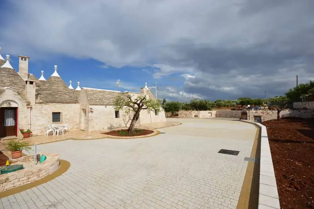 Tradizionali trulli bianchi con tetti conici, ulivi, cortile lastricato, sotto un cielo nuvoloso.