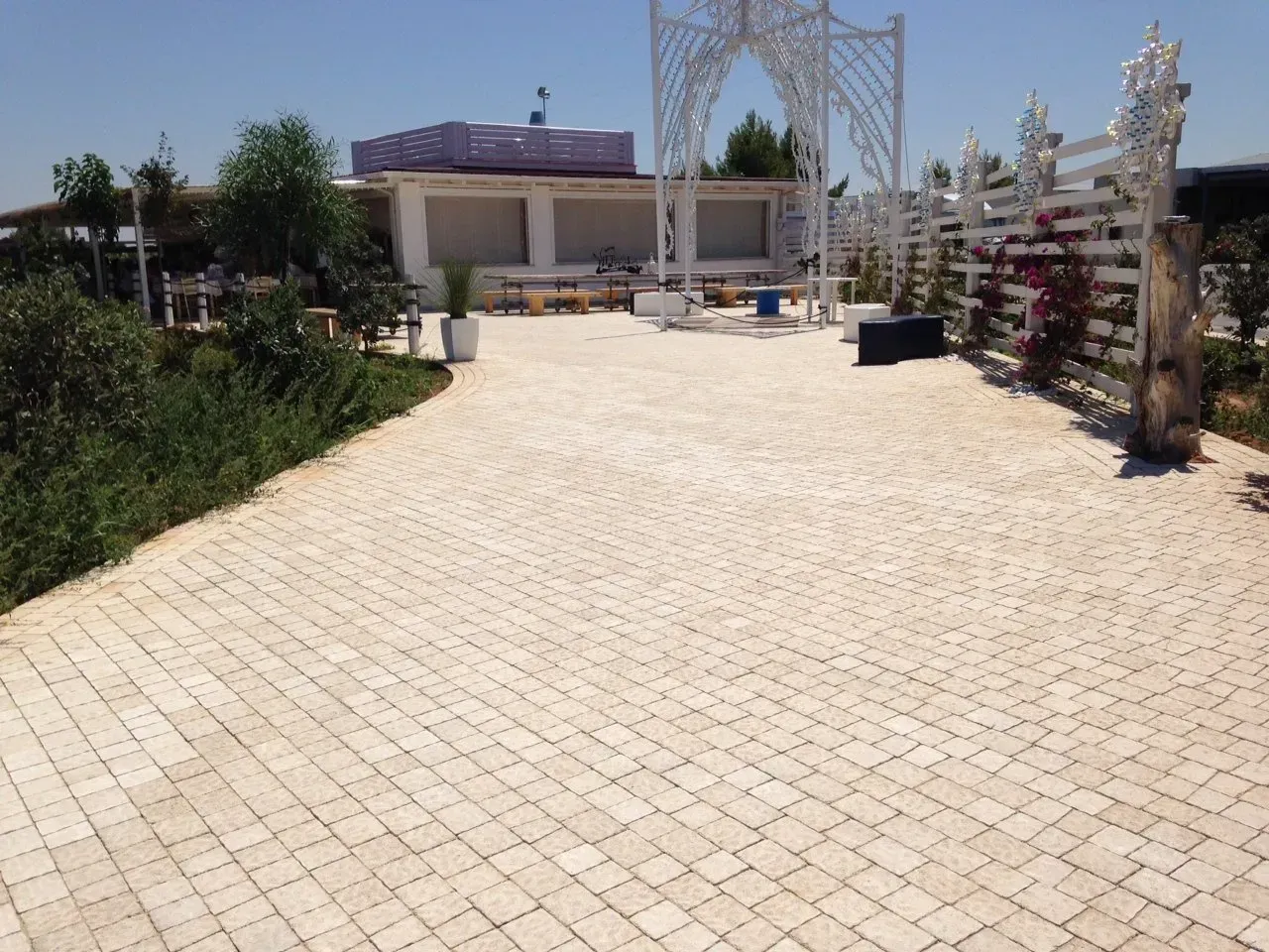 Un cortile lastricato conduce a una struttura bianca con un arco. Verde lussureggiante e cielo azzurro.