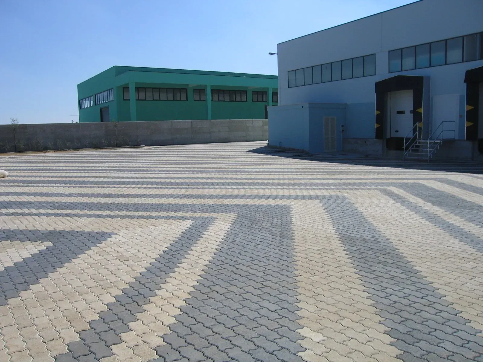 Area pavimentata con mattoni ad incastro grigio chiaro e scuro; sullo sfondo un edificio verde e uno blu.