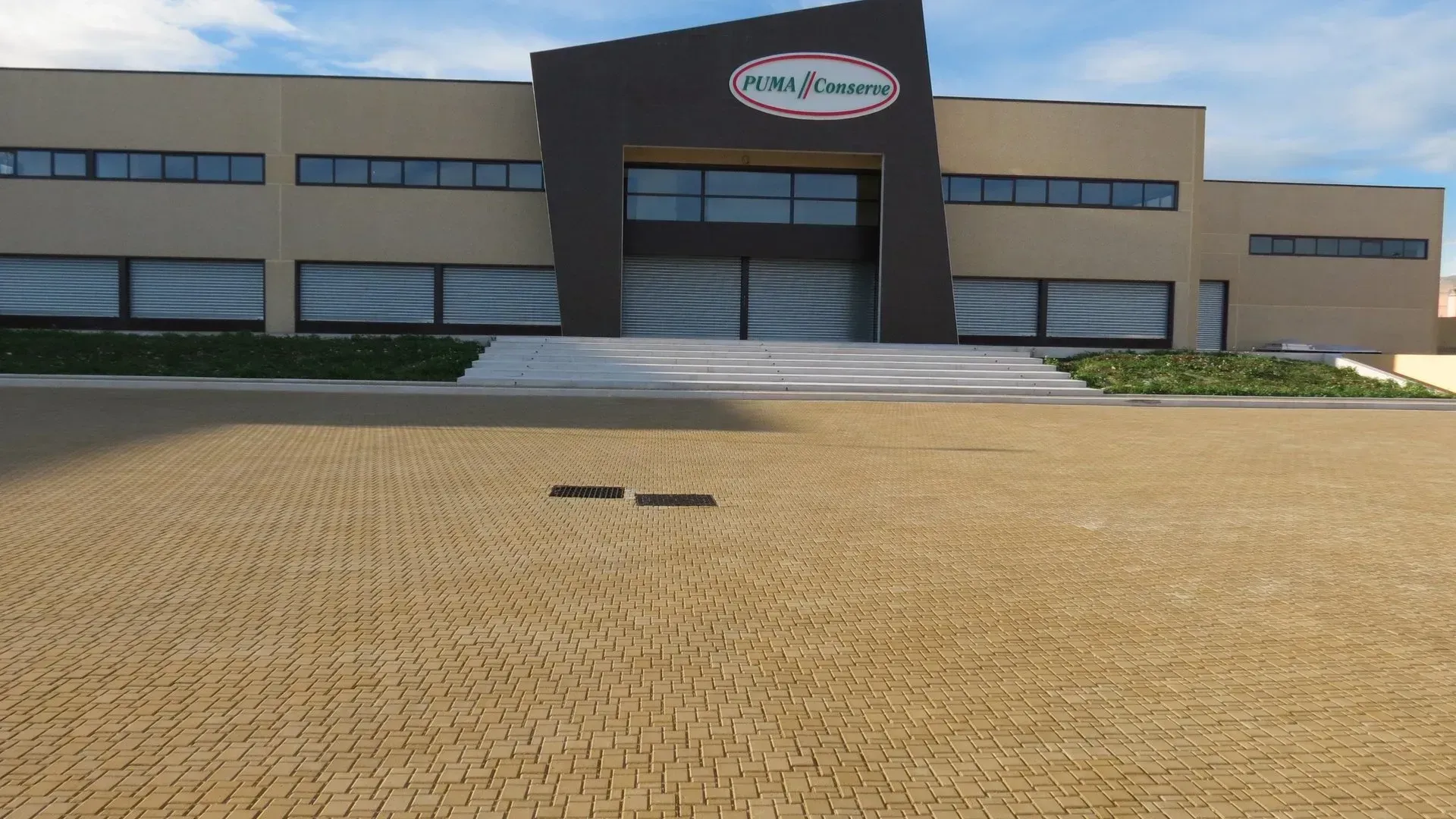 Edificio in mattoni color marrone chiaro con ingresso e logo scuri, situato su un terreno pavimentato in mattoni color marrone chiaro, sotto un cielo azzurro.