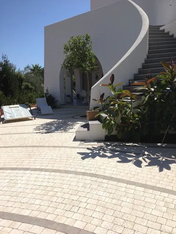 Vista esterna di un edificio bianco con scala curva, patio acciottolato e verde sotto un cielo luminoso.