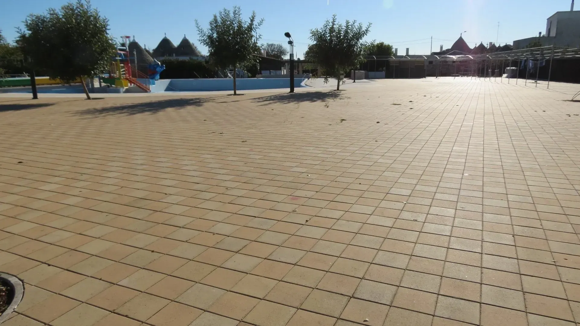 Piazza piastrellata beige con alberi radi, cielo terso e un parco giochi sullo sfondo.