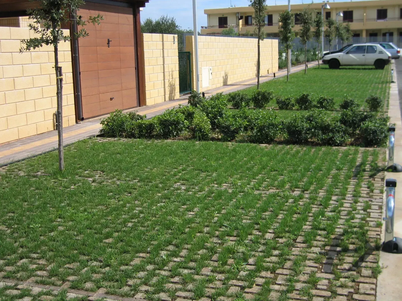 Parcheggio verde con griglia di cemento riempita d'erba, arbusti, alberi e muri color marrone chiaro e marrone. Auto parcheggiate in lontananza.