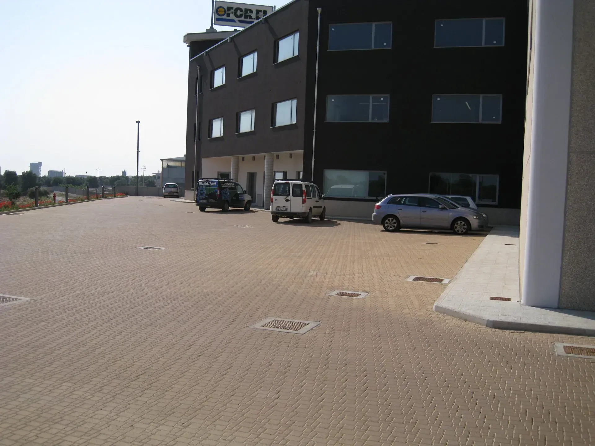 Vista esterna di un edificio di colore scuro con auto parcheggiate su un terreno pavimentato in mattoni.