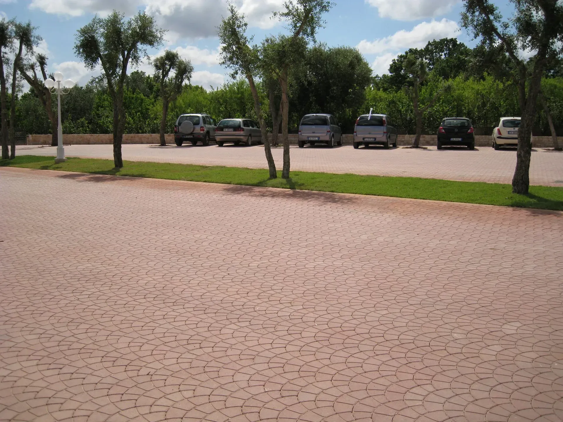Parcheggio con superficie asfaltata rossa, diverse auto parcheggiate, piccoli alberi ed erba verde su uno sfondo di alberi e cielo azzurro.