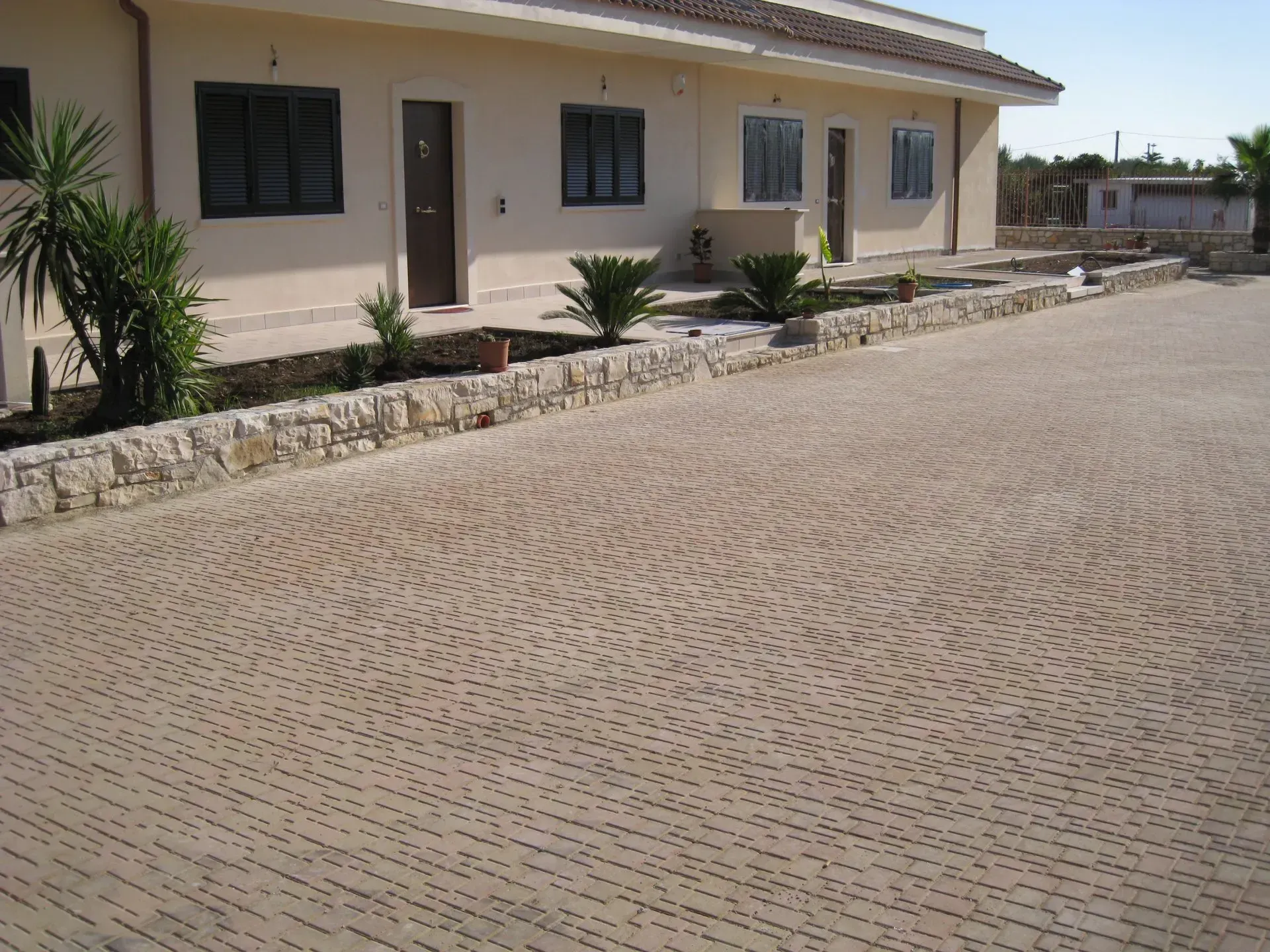 Edificio beige con vialetto decorato, piccoli alberi e bordo in pietra.