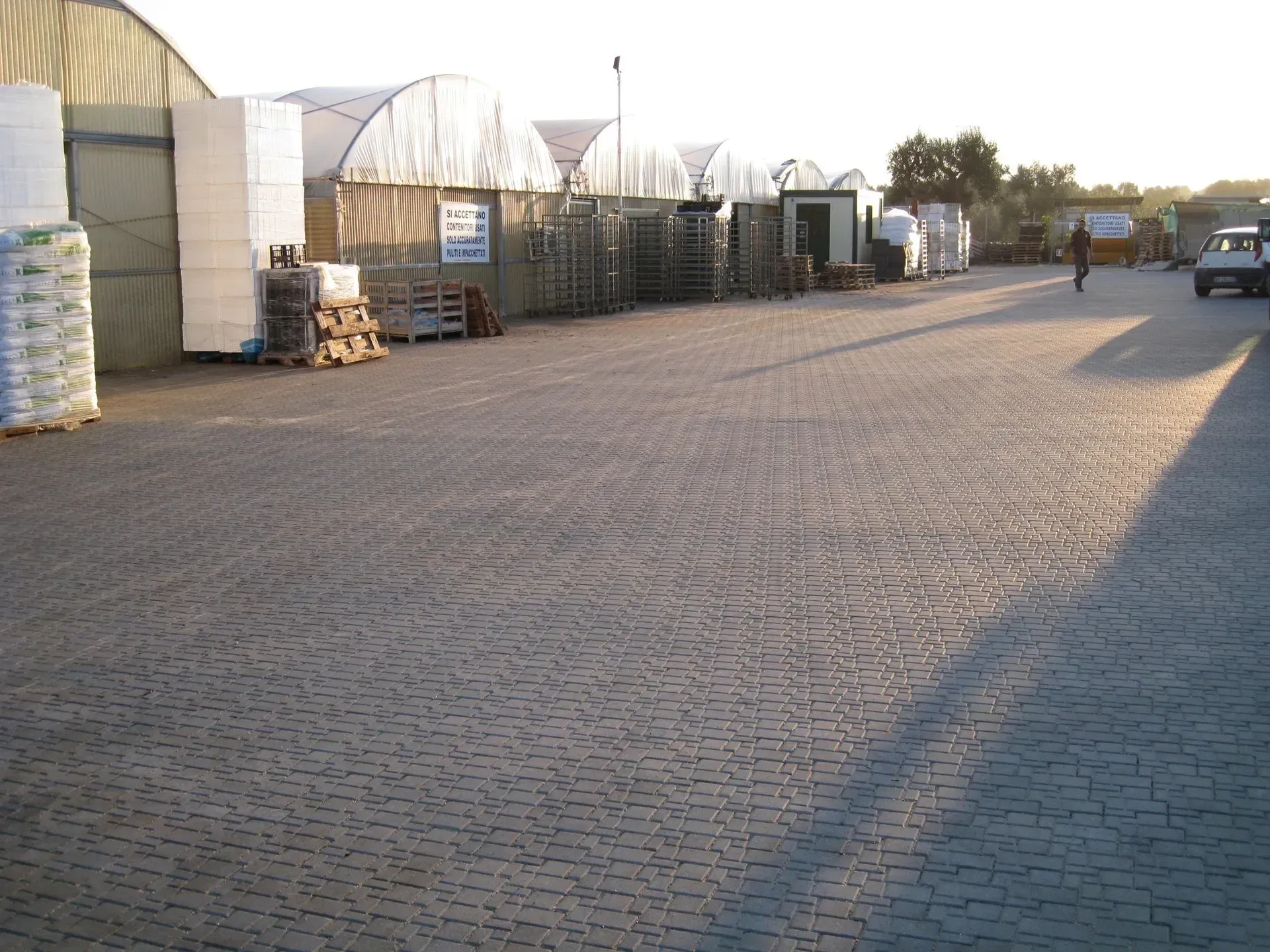 Parcheggio asfaltato con strutture e materiali in stile magazzino. Persona che cammina verso un veicolo sulla destra.