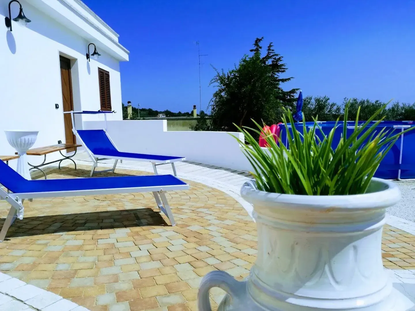 Patio con sedie a sdraio, vaso di piante e una piscina sullo sfondo di un cielo azzurro brillante.