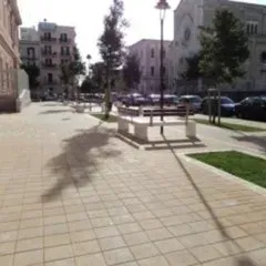 Strada cittadina asfaltata con panchine, alberi, auto parcheggiate ed edifici sullo sfondo in una giornata di sole.