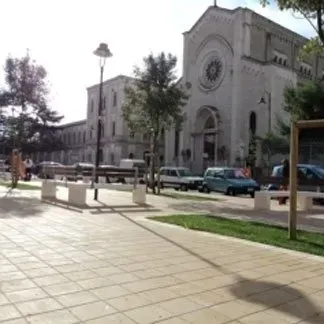 La facciata di una chiesa dai colori chiari, con una grande finestra circolare e un portale ad arco, si affaccia su una strada con auto parcheggiate.