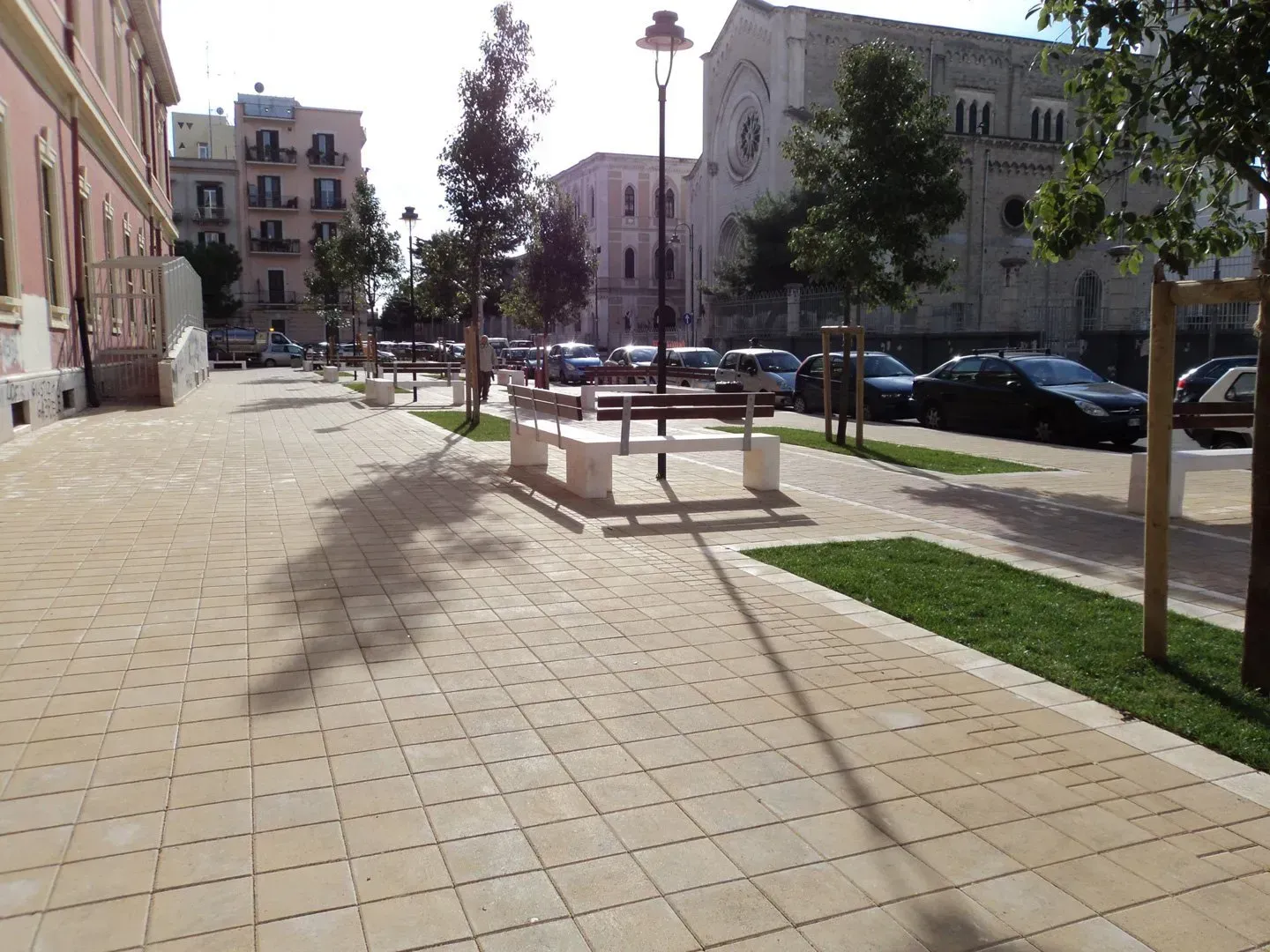 Strada asfaltata con panchine, alberi e auto parcheggiate, di fronte a un edificio con una chiesa sullo sfondo.