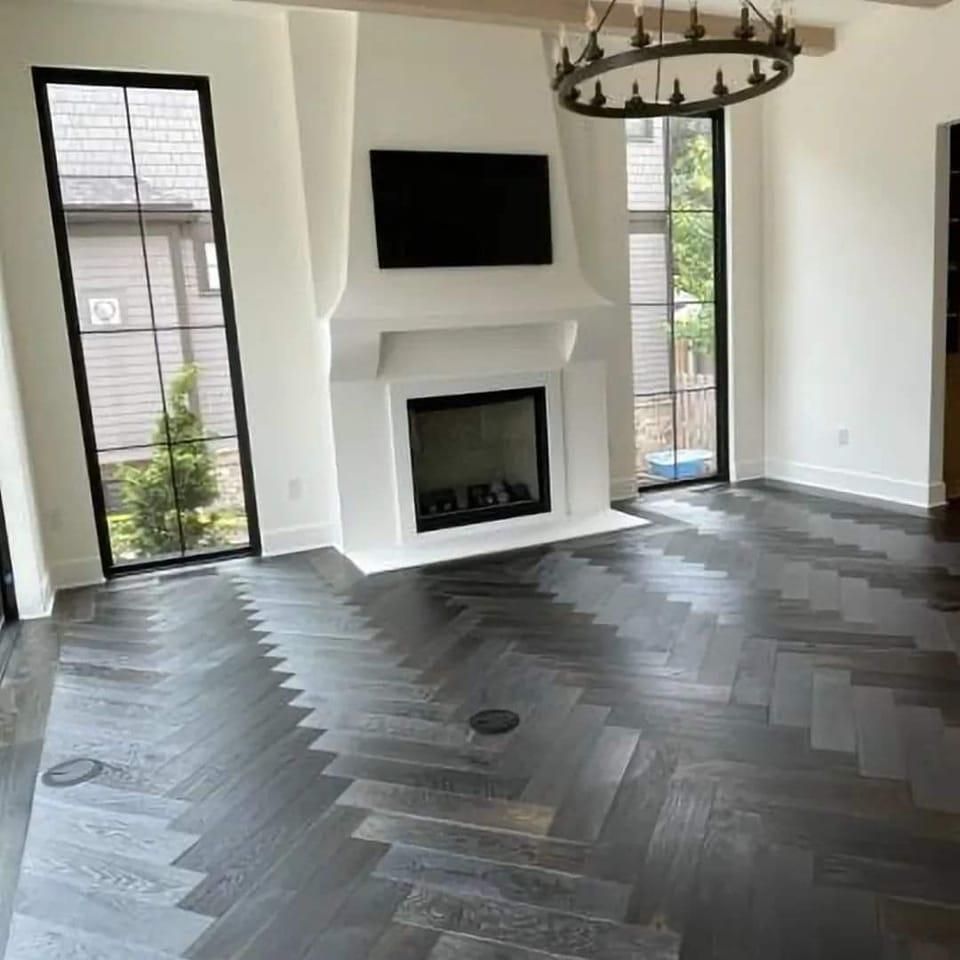 Living room with dark herringbone wood floor, white fireplace, and large windows.
