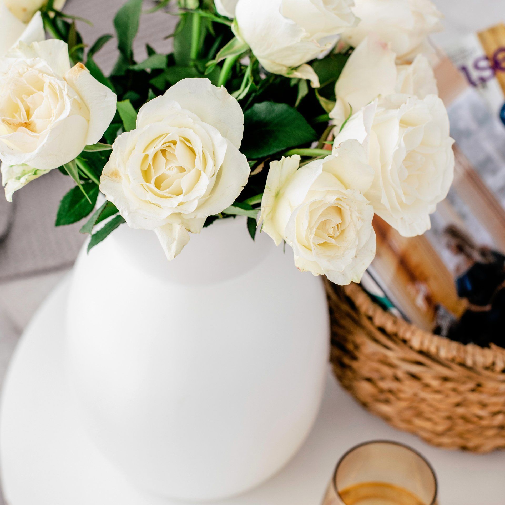 White roses in a white vase on a white table next to a wicker basket and a gold-toned glass.