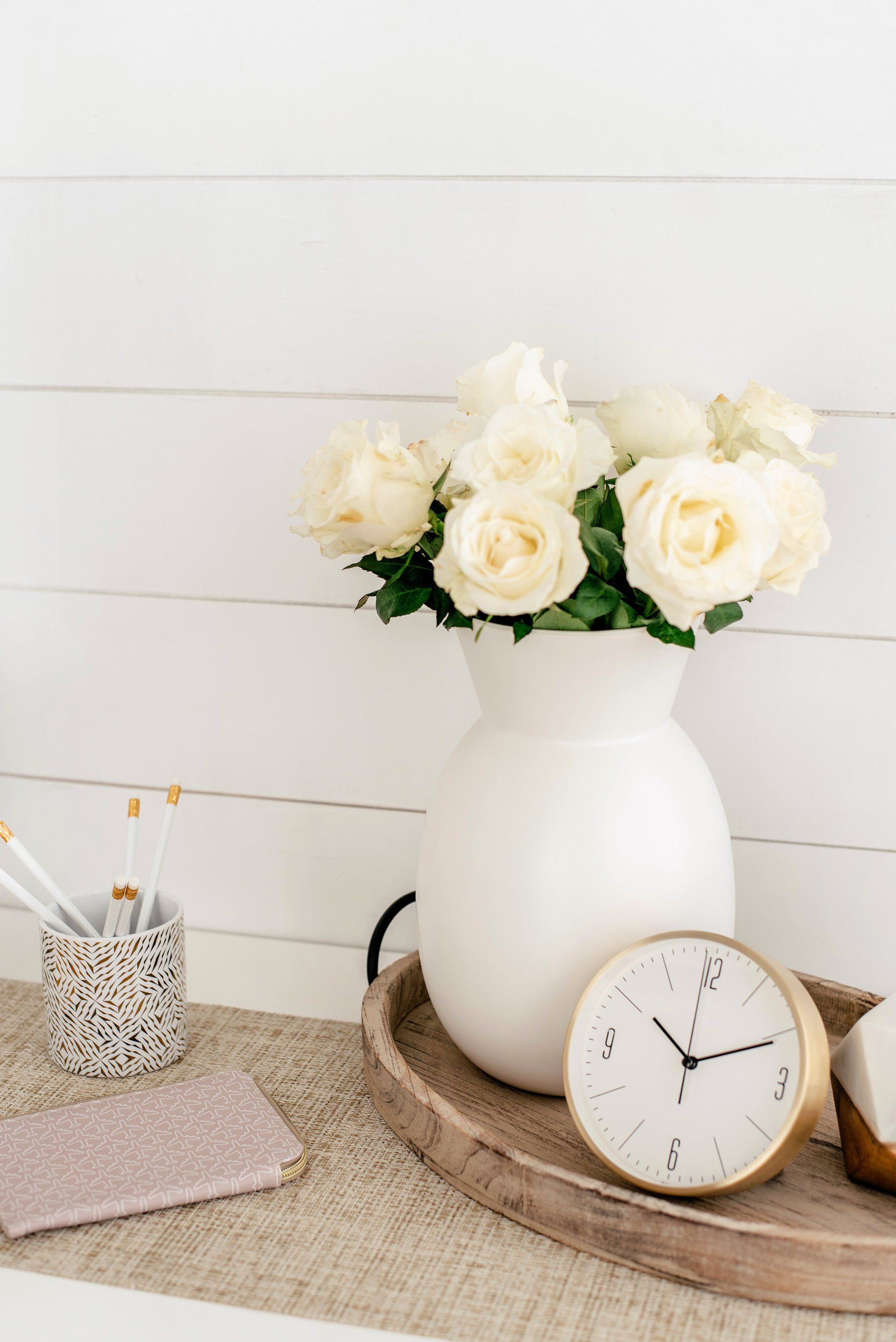 White roses in a vase on a table with a clock and stationery; soft, neutral colors.