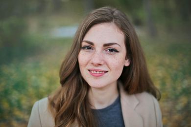 A woman in a tan coat is smiling for the camera in a park.
