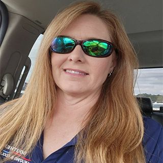 Woman wearing sunglasses in a car, smiling, with long blonde hair, and a blue shirt.