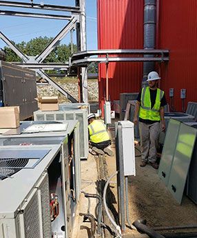 Two workers in hard hats and vests install HVAC units near a red building.
