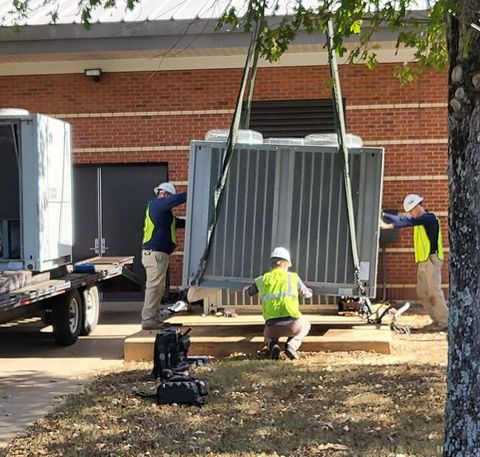 Three workers installing a large electrical transformer with a crane near a brick building; one unloads from a flatbed.