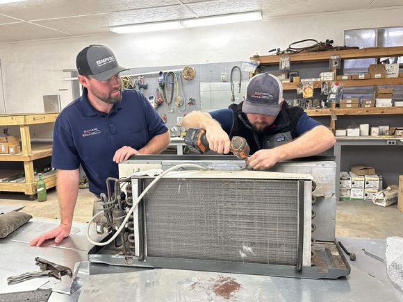Two men working on an air conditioning unit. One uses a drill. Indoors, equipment in the background.