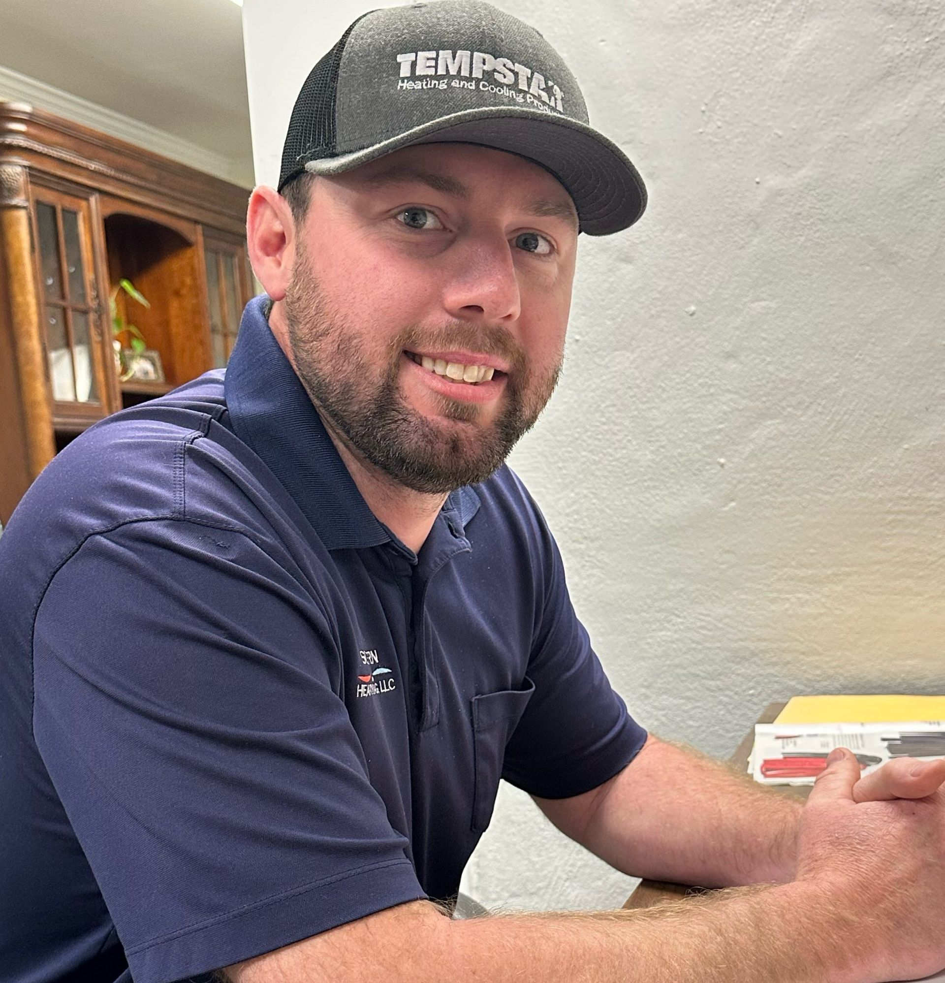 Man wearing Tempstat hat, smiling, seated at a desk, dark blue shirt, neutral wall background.