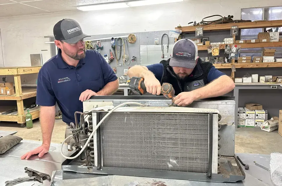 Two men working on an air conditioning unit. One uses a drill. Indoors, equipment in the background.