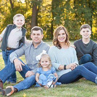 Family sitting on grass smiling; boy in vest, girl with bow, man, woman, teen boy.