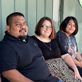 Three people sitting outdoors. Man with a beard, woman with glasses, and a person with short dark hair.