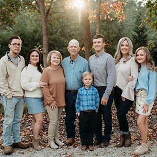 Family of nine poses outdoors, smiling. Includes adults and children, autumn setting, trees and foliage.
