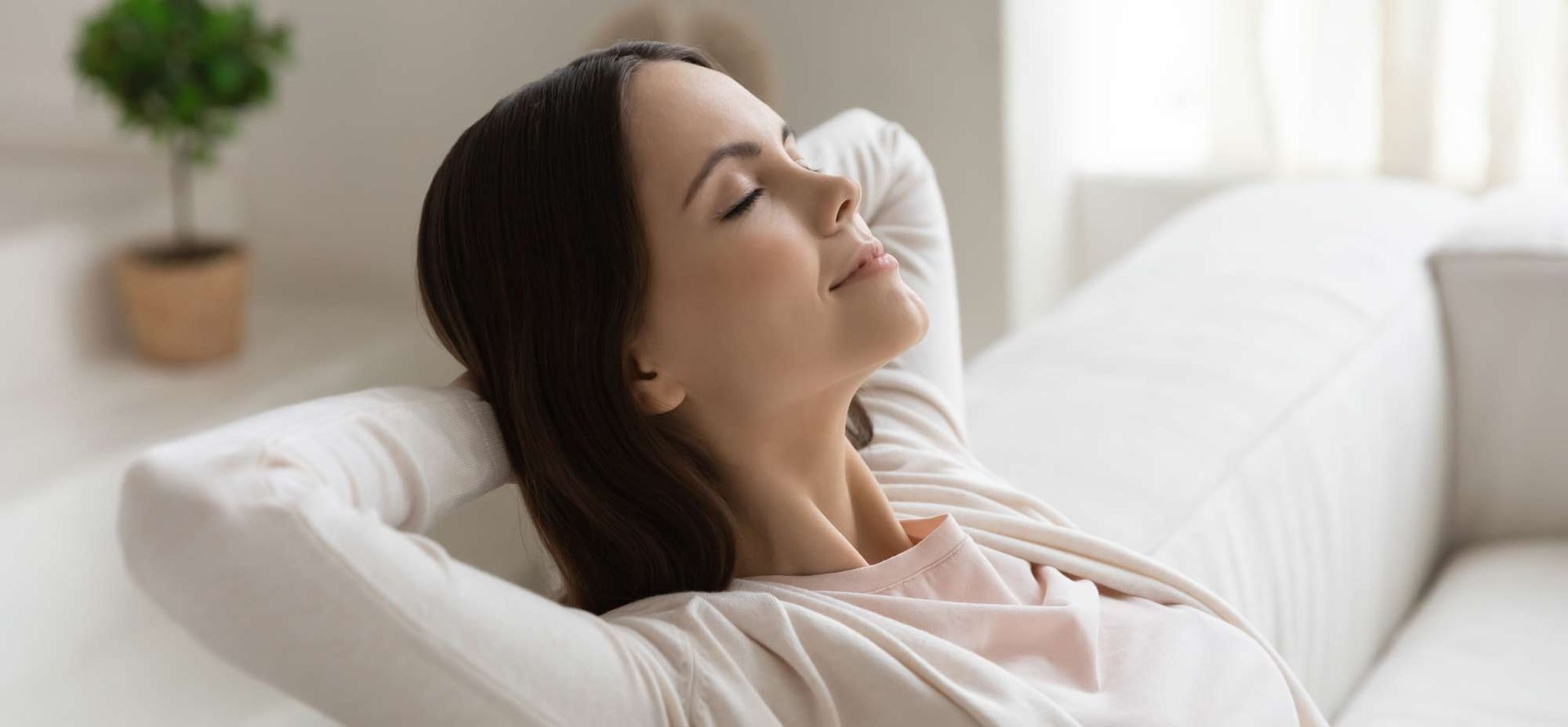 A woman is sitting on a couch with her eyes closed and her hands behind her head.