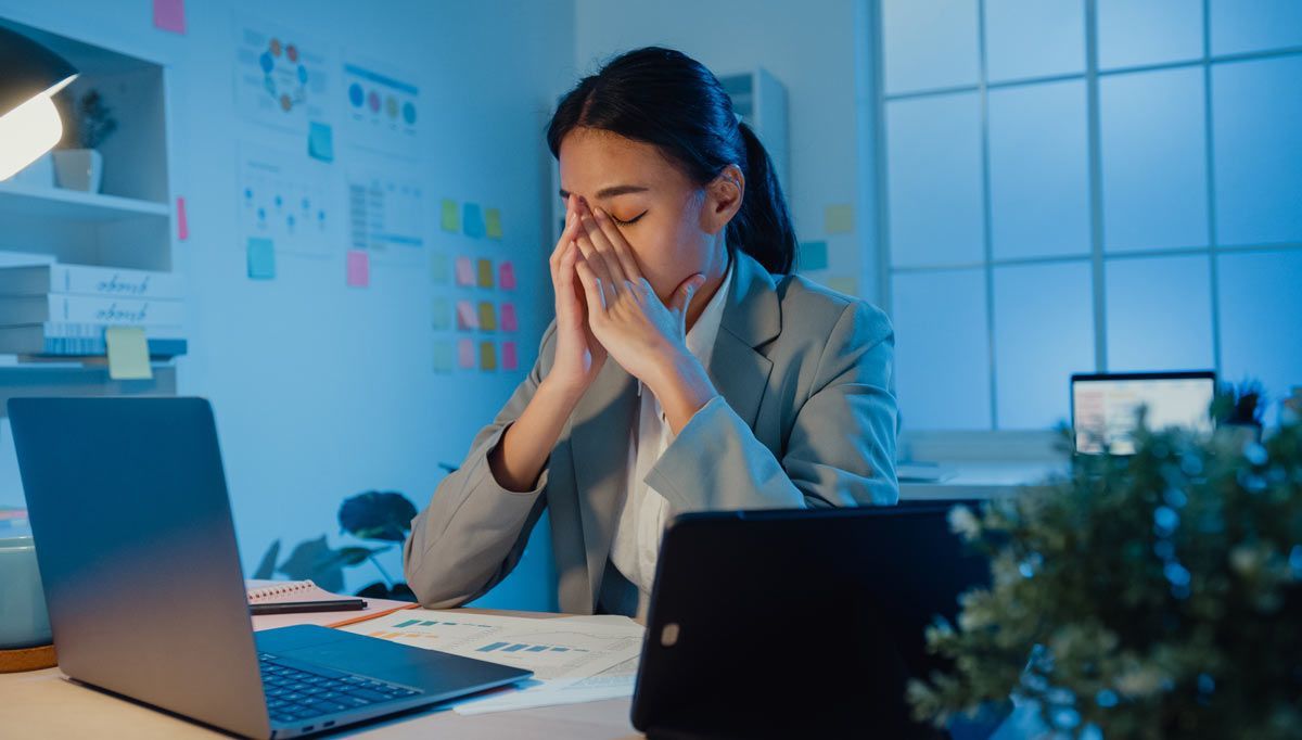 Stressed office worker at a desk with laptop, holding face in hands amid paperwork and plants