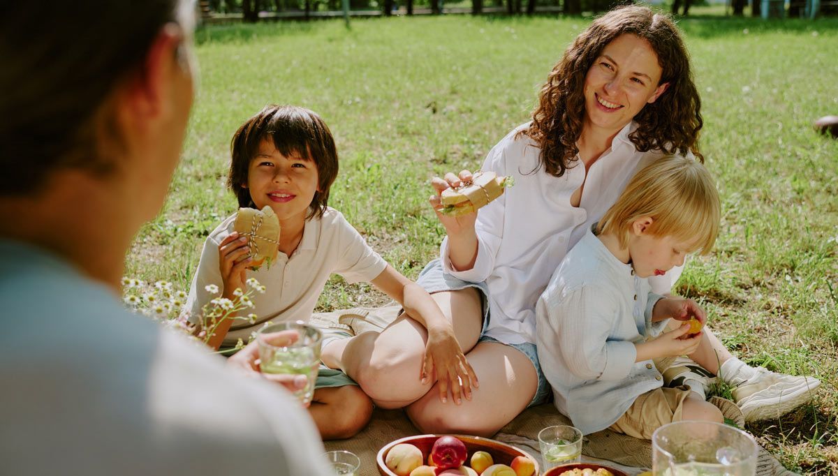 Family picnic on grass, with a woman and two children eating fruit in a sunny park practicing Healthy Summer Weight Management