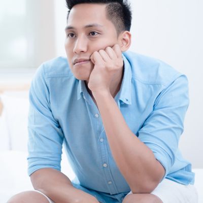 A man in a blue shirt sits on a bed with his hand on his chin, contemplating Testosterone Replacement Therapy NYC by Dr. Dana Cohe,n MD