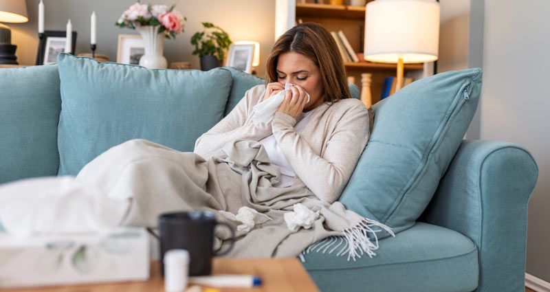 Woman with a cold on a blue couch, blowing her nose into a tissue, wrapped in a blanket unaware of The Surprising Illnesses Associated with a Poorly Functioning Immune System